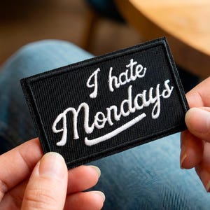 May include: A black rectangular patch with the embroidered phrase "I hate Mondays" in white script. The patch has a black border and is held between two hands.