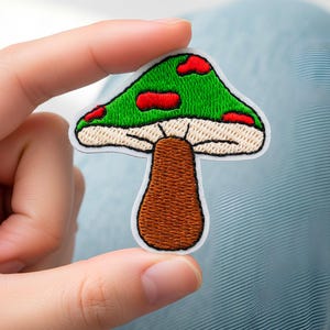 May include: Embroidered mushroom patch with a green cap featuring red spots, a beige stem, and a brown base. The patch has a white border and is held against a light blue background. This patch is ideal for jackets, bags, or other fabric items.