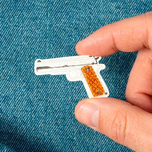 May include: A white and orange embroidered patch in the shape of a handgun. The patch is held against a blue denim background. The handgun has a white body and an orange grip.