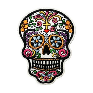 May include: Embroidered patch with a Day of the Dead sugar skull design. The skull is outlined in black and filled with colourful floral and decorative elements in orange, pink, purple, and blue. A cross and heart are centered on the skull.
