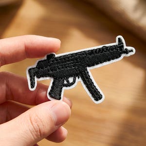 Könnte beinhalten: Schwarzer, gestickter Aufnäher in Form einer Maschinenpistole mit weißem Rand. Der Aufnäher wird von einer Person in der Hand gehalten. Der Aufnäher ist eine detaillierte Darstellung der Schusswaffe.