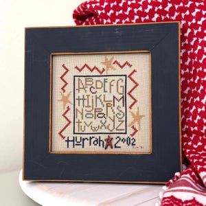May include: A framed cross-stitch sampler with a navy blue wooden frame. The sampler features the alphabet, stars, and the word "Hurrah" with the year 2002. The background is a beige fabric with red and gold stitching.