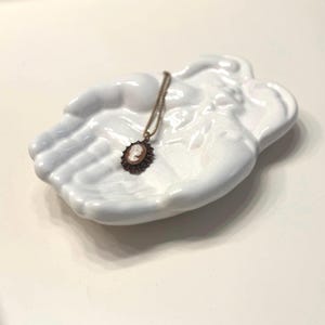 May include: A white ceramic hand-shaped dish holding a cameo pendant necklace. The necklace features a brown cameo pendant with a detailed profile and a dark brown border. The dish is smooth and glossy.