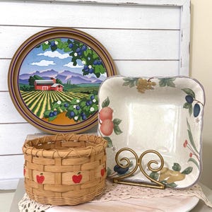 May include: A collection of decorative items, including a round serving tray with a farm scene, a square dish with fruit illustrations, and a small woven basket with red apple designs. A gold-colored napkin holder is also present.