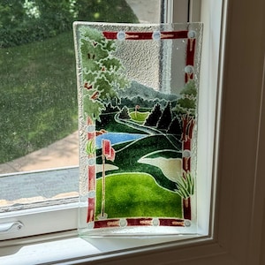 May include: A rectangular glass tray featuring a golf course scene. The artwork depicts a green fairway, blue water, and trees, with red and white accents. The border is decorated with golf balls.