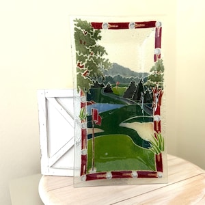 May include: A rectangular glass art piece depicting a golf course scene. The artwork features a green fairway, a pond, and a sand trap. The frame is red with white accents. Trees and a mountain range are in the background.