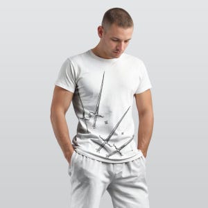 May include: White t-shirt featuring a black illustration of four swords of varying sizes. The swords are arranged diagonally across the shirt, with a few small star-shaped accents. The shirt is worn by a person.