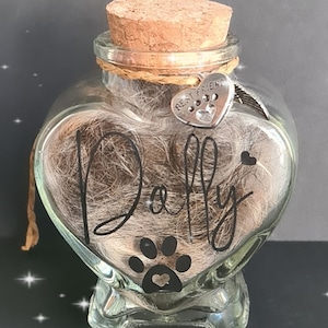 May include: A heart-shaped glass jar with a cork stopper, filled with brown pet fur. The jar has a paw print and the name "Dolly" written on it. A silver charm with the words "Best Friend" and a paw print hangs from the jar.