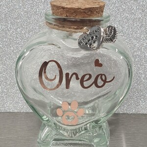 May include: A clear glass heart-shaped jar with a cork lid. The jar has a paw print and the name "Oreo" written in gold lettering. A silver charm with the words "Best Friend" and a paw print hangs from the lid.