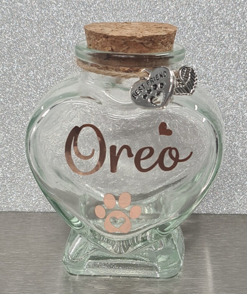 May include: A clear glass heart-shaped jar with a cork lid. The jar has a paw print and the name "Oreo" written in gold lettering. A silver charm with the words "Best Friend" and a paw print hangs from the lid.