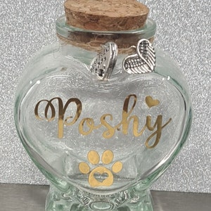May include: A clear glass heart-shaped jar with a cork lid. The jar has a gold paw print with a heart inside and the name "Poshy" written in gold lettering. The jar has two silver charms attached to the lid, one with the words "Best Friend" and the other with angel wings.