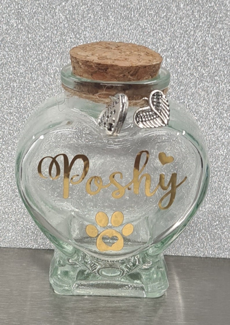 May include: A clear glass heart-shaped jar with a cork lid. The jar has a gold paw print with a heart inside and the name "Poshy" written in gold lettering. The jar has two silver charms attached to the lid, one with the words "Best Friend" and the other with angel wings.