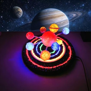 May include: A solar system model featuring a central red sun and illuminated planets in various colours. The base has concentric rings of blue, green, and red lights. The background shows planets and stars.
