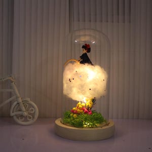 Könnte beinhalten: Dekorative Glaskuppel mit einer Miniaturfigur auf einer Watte-Wolke, beleuchtet von einem warmen Licht. Die Basis enthält Moos und Blumen. Ein Miniatur-weißes Fahrrad mit einem Korb befindet sich im Hintergrund.
