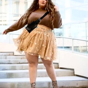 May include: A woman wearing a brown sweater, a black fanny pack, and a beige tulle skirt with gold stars. She is standing on a set of stairs.