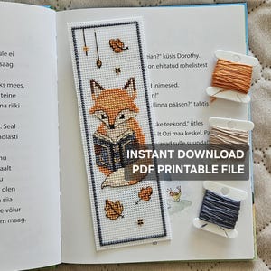 May include: A rectangular cross-stitch bookmark on white Aida cloth featuring a fox reading a book, surrounded by falling autumn leaves. Three plastic bobbins with orange, beige, and dark gray embroidery floss are placed to the right.