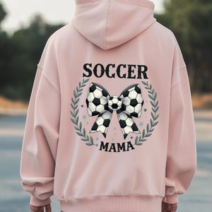 May include: A light pink hoodie with a soccer-themed design. The back features a bow made of soccer balls, surrounded by a green wreath, with the words "SOCCER MAMA" in black lettering.