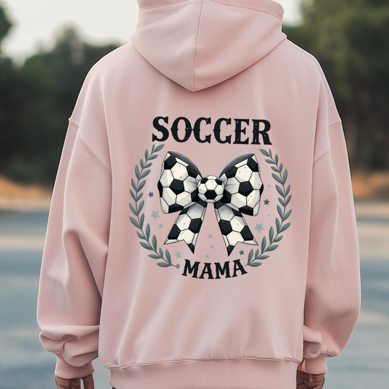 May include: A light pink hoodie with a soccer-themed design. The back features a bow made of soccer balls, surrounded by a green wreath, with the words "SOCCER MAMA" in black lettering.