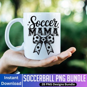 May include: White ceramic mug with the words "Soccer MAMA" in black script and block letters, with a soccer ball bow design. The mug is held in a hand against a blurred green background.