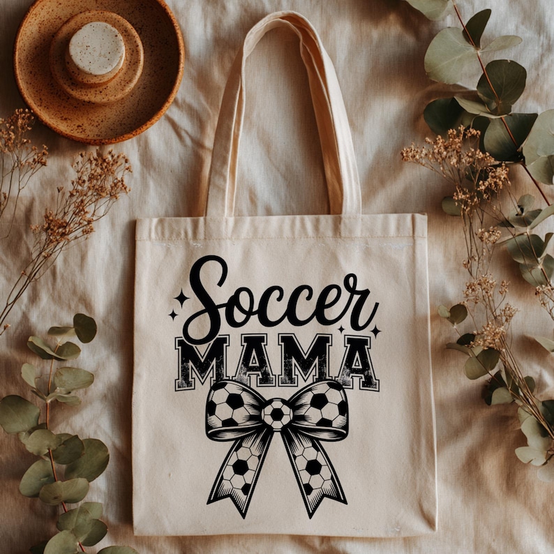 May include: Beige tote bag with the words "Soccer MAMA" in black script and block letters. A black and white soccer ball bow design is below the text. The bag has two handles and is made of a canvas-like material.