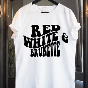 May include: White t-shirt with the words "RED WHITE & BRUNETTE" in large, bold, black, retro-style lettering. The shirt is hanging on a black hanger. The background is a neutral color.