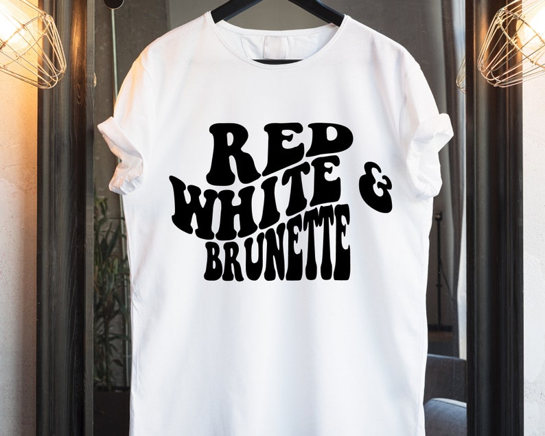 May include: White t-shirt with the words "RED WHITE & BRUNETTE" in large, bold, black, retro-style lettering. The shirt is hanging on a black hanger. The background is a neutral color.
