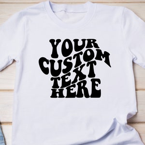 May include: White t-shirt with the text "YOUR CUSTOM TEXT HERE" in a black, retro-style font. The shirt is made of a soft material and has short sleeves.