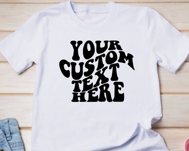 May include: White t-shirt with the text "YOUR CUSTOM TEXT HERE" in a black, retro-style font. The shirt is made of a soft material and has short sleeves.