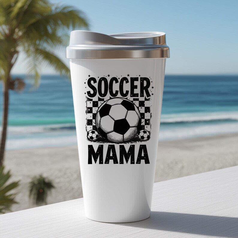 May include: White travel mug with a silver lid featuring the text "SOCCER MAMA" in black. The design includes a soccer ball graphic with a checkered background and small soccer balls. The background shows a beach and ocean.