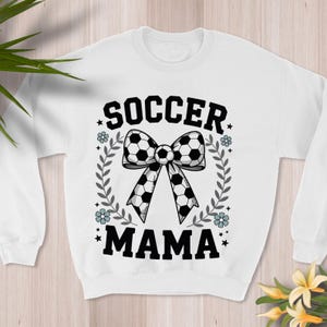 May include: White sweatshirt with the words "SOCCER MAMA" in black block letters. A bow made of soccer balls is in the center, surrounded by a wreath of leaves and flowers. The design is centered on the front of the sweatshirt.