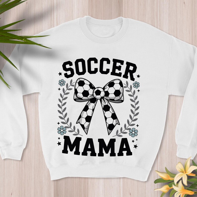 May include: White sweatshirt with the words "SOCCER MAMA" in black block letters. A bow made of soccer balls is in the center, surrounded by a wreath of leaves and flowers. The design is centered on the front of the sweatshirt.