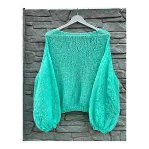 May include: A turquoise knitted sweater with a wide neckline and voluminous sleeves. The sweater is displayed on a white hanger against a grey stone wall. The knitwear has a loose, airy texture.