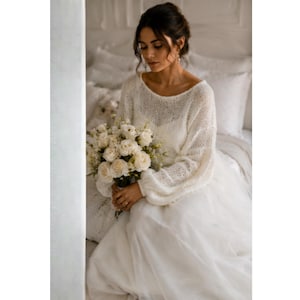 May include: A bride in a white wedding dress with a long skirt and a knitted, off-the-shoulder sweater. She holds a bouquet of white roses. The setting is a softly lit room, creating a romantic atmosphere.