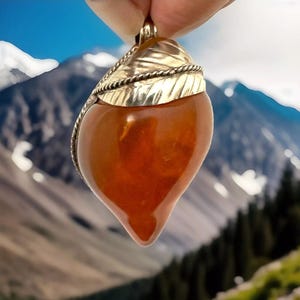 May include: A teardrop-shaped pendant with a warm amber hue. The pendant is topped with a gold-colored leaf design and a gold-colored bail. The background features a blurred mountain landscape.