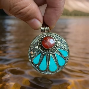 May include: A circular silver pendant featuring a central red stone and turquoise teardrop-shaped inlays. The pendant has an intricate design and is held by a hand.