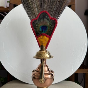 Könnte beinhalten: Dekorativer Kupfer- und Messing-Räuchergefäß mit fächerförmiger Spitze aus dunkelbraunen Federn. Der Fächer hat einen roten und gelben Akzent mit einer Pfauenfeder in der Mitte.