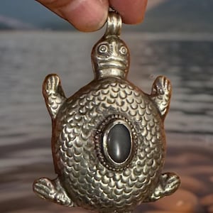 May include: A silver-toned metal pendant shaped like a turtle with a textured shell pattern and a smooth, dark oval stone set in the center. The pendant features a small loop at the top for attachment and has a detailed head, four flippers, and a tail.