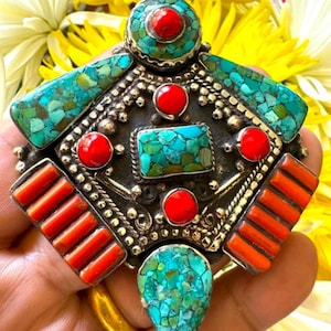 May include: A detailed pendant with turquoise and red stone accents. The silver-toned metal pendant features a central rectangular turquoise stone, surrounded by red and turquoise stones. The pendant is set against a backdrop of yellow and white flowers.