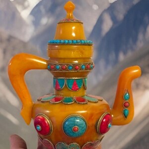 May include: Ornate amber-colored teapot with turquoise and red accents. The teapot features a decorative lid and handle, with colorful circular embellishments. The background shows a mountain range.