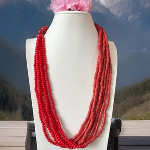 Könnte beinhalten: Eine mehrreihige Halskette aus roten Korallenperlen. Die Halskette wird auf einer weißen Mannequin-Büste vor dem Hintergrund einer schneebedeckten Bergkette präsentiert.
