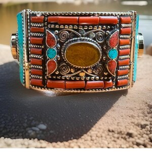 May include: A rectangular bracelet with a silver-colored metal frame. The bracelet features turquoise, red, and yellow-brown stones. The design includes a central oval stone and smaller stones in a symmetrical pattern. The bracelet is displayed on a sandy surface.