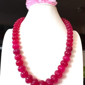 Op de afbeelding: Een levendige, robijnrode kralenketting, gepresenteerd op een witte mannequin. De ketting heeft grote, gesneden kralen met een getextureerd oppervlak. Een roze bloem siert de bovenkant van de mannequin. Een opvallend sieraad.