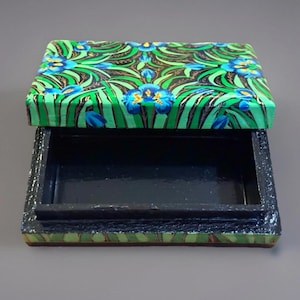 May include: An open, rectangular box with a lid. The lid is decorated with a green and blue floral pattern. The box itself is black with a textured, green and brown border.