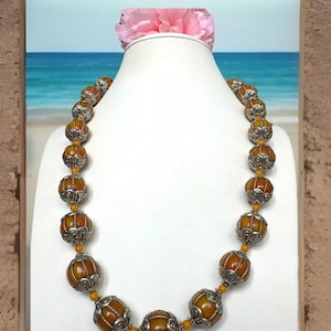 May include: A necklace made of amber beads with silver accents. The beads are round and have a warm, honey-like color. The necklace is displayed on a white mannequin against a backdrop of a beach scene.