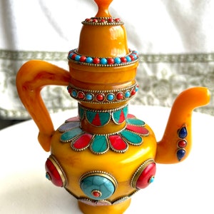 May include: A small yellow teapot with a lid decorated with turquoise, red, and gold accents. The teapot has a curved spout and handle and is adorned with colorful gemstones.