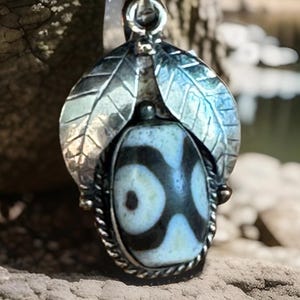 May include: A silver pendant with a leaf design and an oval-shaped stone. The stone is white with black circular patterns. The pendant hangs from a white ribbon. The background is blurred.