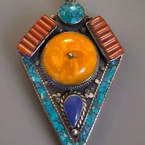 May include: A silver pendant with a large, round amber stone at the center. The pendant features turquoise, coral, and a blue gemstone, set in a geometric design. The pendant is approximately 2 inches in length.