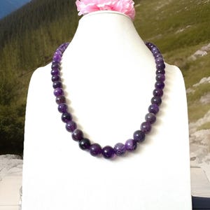May include: A necklace of polished, round, purple amethyst beads displayed on a white bust form. A pink flower sits atop the form. The background features a mountain landscape with a blue sky.