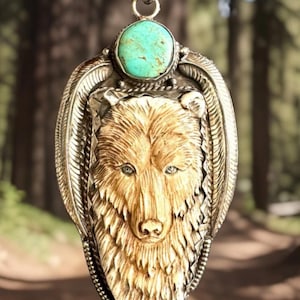 May include: A silver pendant featuring a carved bear head in a light tan material, framed by an ornate silver border with feather-like engravings. A round, polished turquoise stone is set at the top, below a decorative silver bail.