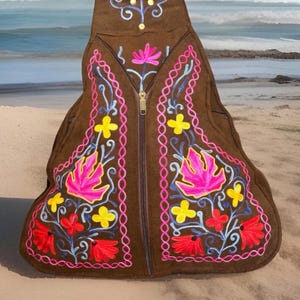 May include: Brown suede backpack with colourful floral embroidery. The design features pink, red, and yellow flowers with blue accents. The bag has a zip closure and is set against a beach backdrop.